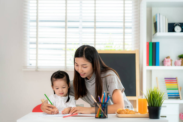 Asiatische Kindergartenschülerin mit Mutter malt Bild in Buch mit Farbstift zu Hause, Homeschooling und Fernstudium - Foto, Bild