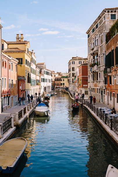 Classic view of the narrow sea Venetian canal. Colorful buildings stand opposite each other, boats moored near shore, gondolier rides tourists on a gondola in Venice, Italy. Blue skies on a sunny day. - Foto, afbeelding