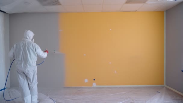 Worker wearing white workwear painting the orange wall in white color by airless spray gun. Airless Spray Painting. Repair at home or office. - 映像、動画