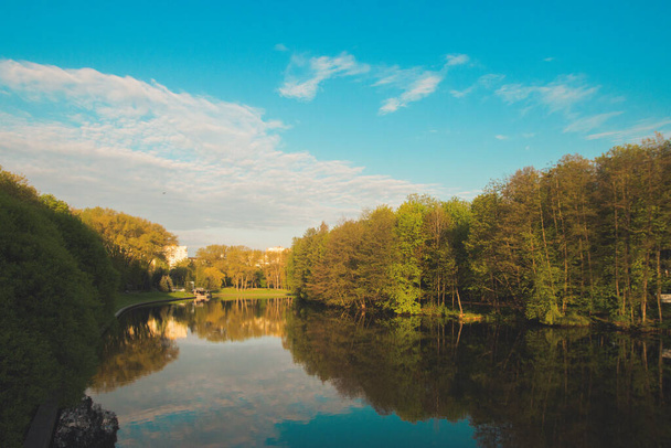 Sabahın erken saatlerinde şehir parkında bir göl. Sessiz ve huzurlu, uyum içinde, insan yok. Taze yeşil ağaçlar ve çimenler, mavi gökyüzü ve suya yansıyan bulutlar. Ilık güneş ışığı. Minsk Belarus. - Fotoğraf, Görsel