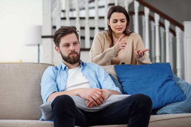 Confused man sits on the couch while his girlfriend yells and quarrels with him at home. Big family problems. - Valokuva, kuva