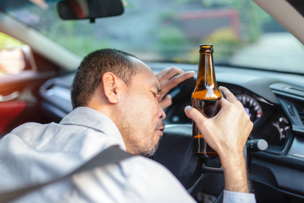 Drunk asian young man drives a car with a bottle of beer with sunset background, Dangerous driving concept - Photo, Image