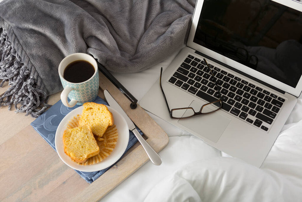 Trabajando desde casa concepto con portátil, taza de café, en la cama rodeada por una manta con un muffin de maíz y vasos. Una vista superior elegante espacio de trabajo moderno
 - Foto, imagen