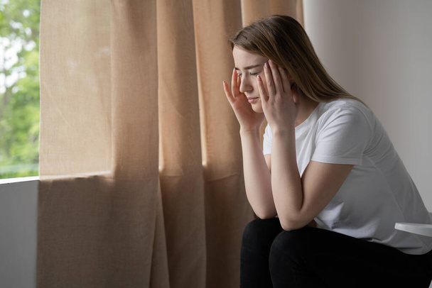 depressed woman sitting on a couch at home by the window. Headache after overwork. Young blond girl suffering from migraine pain and bad mood - Photo, Image