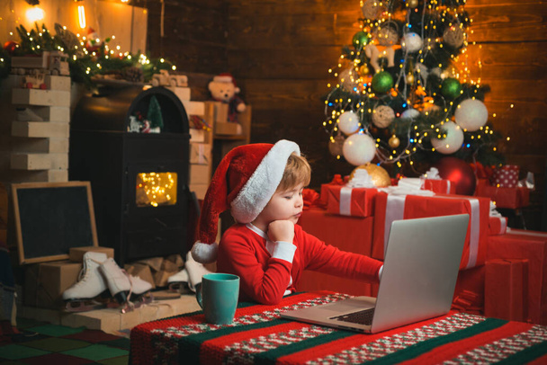 Wish List for Santa. Child type letter to Santa. On-line Christmas shopping for kids. Little kid is wearing Santa clothes sitting by his laptop. Christmas concept. - Valokuva, kuva