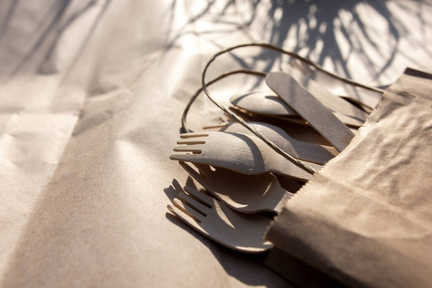 eco-friendly wooden forks, spoons and knives in a craft bag. biodegradable disposable tableware. shadows from the grass. picnic appliances on the side. - Photo, Image