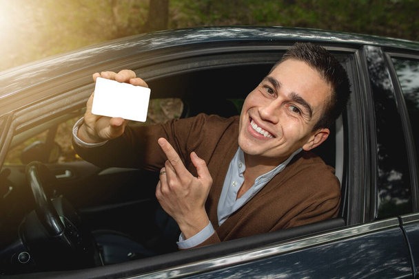 Retrato de joven caucasiano sonriente dentro de su coche nuevo publicado fuera de la ventana. Poner el dedo en la licencia de conducir u otro documento, espacio libre para el texto en la tarjeta.. - Foto, imagen
