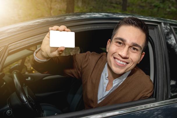 Πορτρέτο του νέου χαμογελαστού άνδρα μέσα στο νέο του αυτοκίνητο δημοσιεύτηκε έξω από το παράθυρο που δείχνει την άδεια οδήγησης ή άλλο έγγραφο, ελεύθερος χώρος για κείμενο στην κάρτα. - Φωτογραφία, εικόνα