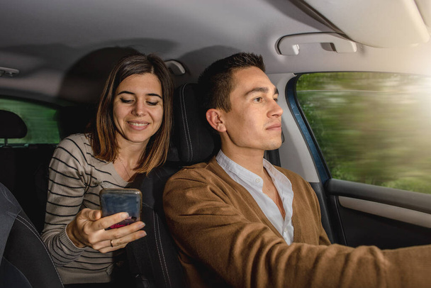 Caucasian couple inside car. Man driving and woman holding smartphone to write emails, select music or app navigation maps. Moving car. - Fotó, kép