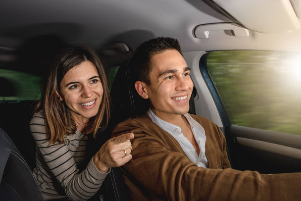 Caucasian couple inside car smiling. Woman is pointing something outside with finger while man is driving. Happy faces in a journey or travel. Moving car with blurred background. - Foto, Imagem