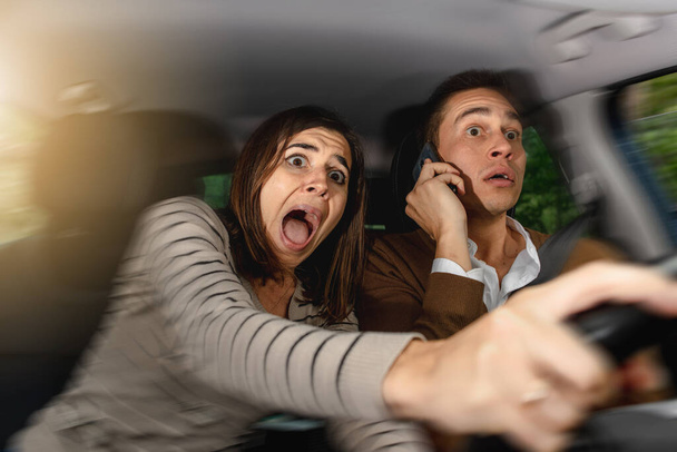 Φοβισμένο ζευγάρι μέσα στο αυτοκίνητο. Ο οδηγός μιλάει στο τηλέφωνο ενώ ο επιβάτης αρπάζει το τιμόνι προσπαθώντας να αποφύγει το εμπόδιο ουρλιάζοντας συντριβή. Επικίνδυνη οδήγηση. Παραβίαση κανόνων κυκλοφορίας και νόμων. Οδήγηση - Φωτογραφία, εικόνα