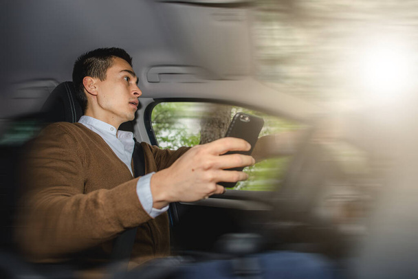 Young man is talking on the phone while driving fast. Scared because distracted by the conversation is causing a car accident. Violation traffic rules and laws. Blarred background because high speed. - Photo, Image