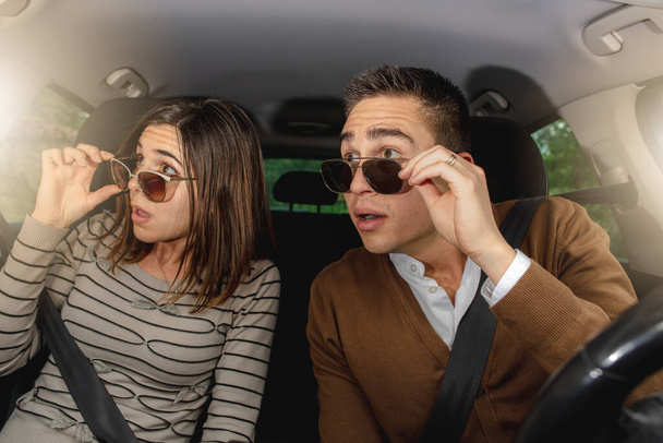 Καυκάσιο ζευγάρι μέσα σε αυτοκίνητο σε δράση, οδήγηση. Βγάλτε τα γυαλιά ηλίου για να δείτε κάτι καλύτερο στο δρόμο κατά τη διάρκεια του ταξιδιού. Έκπληκτος. - Φωτογραφία, εικόνα