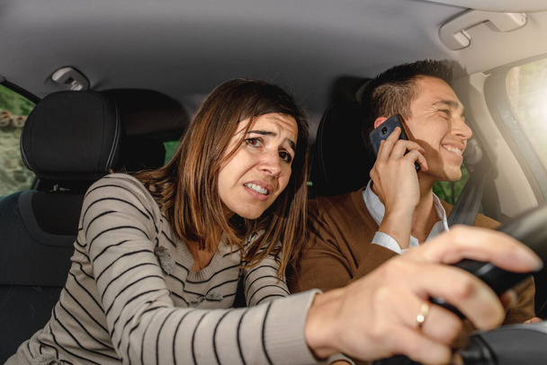 Young man talking on the phone while driving and woman passenger grabs steering wheel trying to avoid an obstacle. Scared and screaming couple is getting a car accident. - Photo, Image