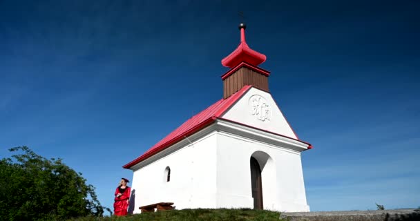 Θλιβερή γυναίκα με κόκκινο, ιστορικό βικτοριανό φόρεμα με καπέλο που περπατάει από την εκκλησία. Μικρό λευκό εκκλησάκι. - Πλάνα, βίντεο