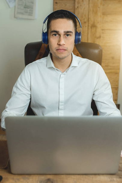Függőleges portré a munkahelyén ülő fiatalemberről, aki az asztal előtt ül, és a laptop fejhallgatót visel. Munkahelyi, munkáltatói és ügyfélszolgálati koncepció. - Fotó, kép