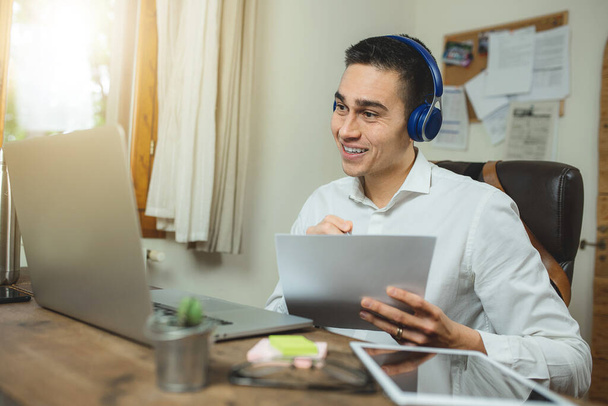 Young businessman having a work video call with headphones. Workplace with laptop, paper, post it, tablet and other personal objects in the wooden office desk. Call center, employer concept - Photo, Image