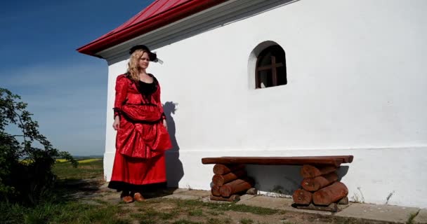 Blond lady in red costume dress sitting in nature near chapel relaxing - Footage, Video