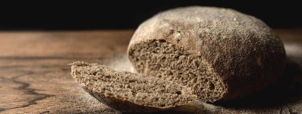 Close up of black rye loaf of sliced bread on wooden table with flour. Dark background. - Photo, Image