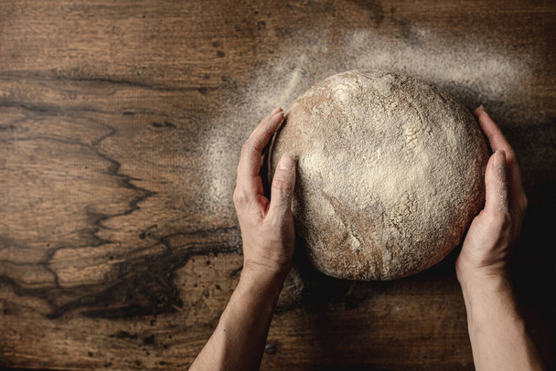 Vista superior das mãos da mulher em torno de um pedaço de pão integral de cereais. Farinha na mesa de madeira e espaço livre para texto à esquerda. - Foto, Imagem