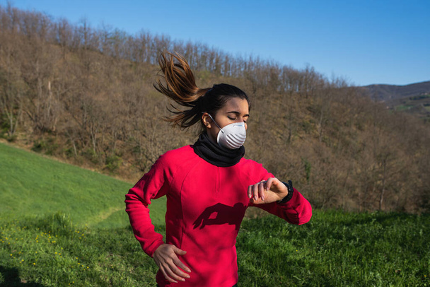 Sportieve jonge vrouw draagt gezichtsmasker van een coronavirus pandemie. Kijken naar het polshorloge na lichaamsbeweging, fitness en hardlopen in de buitenlucht. - Foto, afbeelding