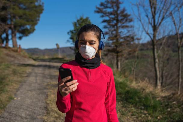 Kulaklıklı ve akıllı telefonlu sportif bir kadın açık havada Coronavirus sırasında yüz koruyucu maskeyle spor yapıyor. Salgın, sağlıklı yaşam tarzı, spor anlayışı. - Fotoğraf, Görsel
