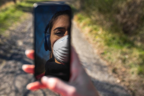 Πορτρέτο νεαρή γυναίκα με προστατευτική μάσκα προσώπου κάνει αθλητισμό εξωτερική. Κρατώντας ένα smartphone και εικόνα που αντανακλάται στην οθόνη. - Φωτογραφία, εικόνα