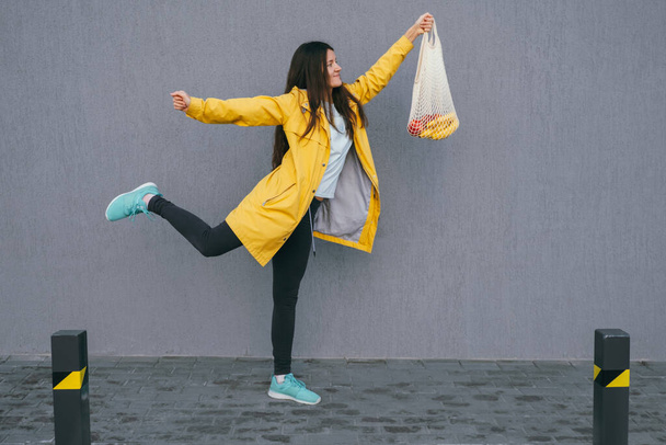 active young woman in a yellow raincoat carrying string bag with fresh bananas and apples on the street against gray wall ,copy space - Foto, immagini
