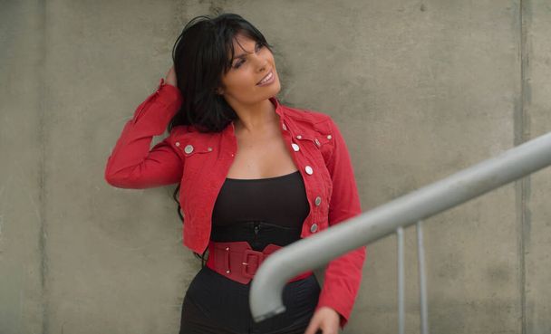 Bella donna con i capelli lunghi in giacca rossa in piedi vicino al muro di cemento e sorridente. Persone reali in città
 - Foto, immagini