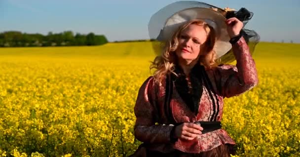 Blond noblewoman with large hat in the middle of oilseed rape yellow field in historical costume dress. - Footage, Video