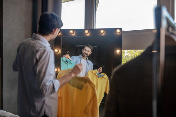 Man reflecting in the mirror with tshirts in his hands - Foto, imagen