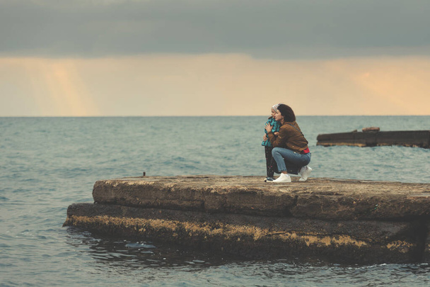 Äiti ja tytär seisovat aallonmurtajalla meren rannalla ja katsovat horisonttia auringonlaskun aikaan. Loma meren aktiivinen kävelee ja terveellistä elämäntapaa. Vaakasuuntainen suunta ja kopiotila
. - Valokuva, kuva