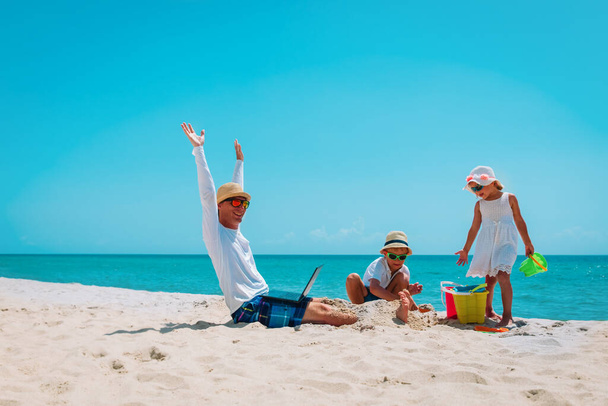 ノートパソコンで仕事をしようとする幸せな父親とビーチで砂と遊ぶ子供たち - 写真・画像