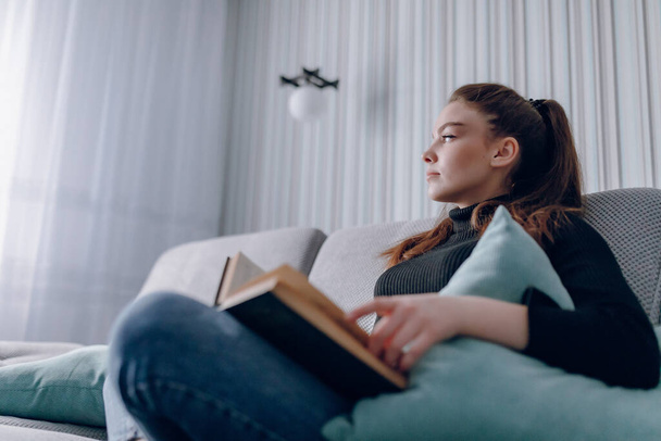 νεαρή ελκυστική κοπέλα στον καναπέ διαβάζει ένα χάρτινο βιβλίο. διανοητική ανάπτυξη. χρήσιμη χρήση του χρόνου στο σπίτι. ζεστή άνεση στο σπίτι. - Φωτογραφία, εικόνα