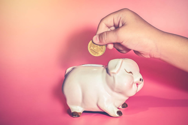 La mano del niño deja caer una moneda en Piggy Bank para ahorrar con una pila de monedas en la mesa en casa, ahorre dinero para el aprendizaje futuro
 - Foto, Imagen