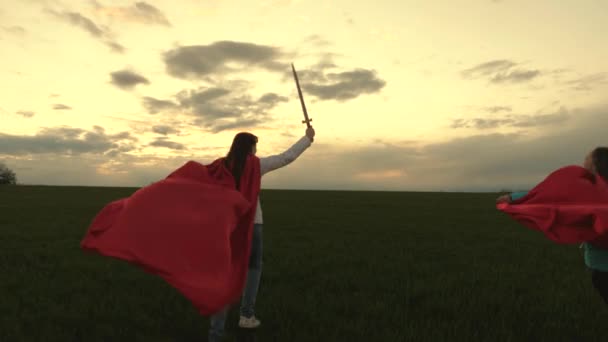 free young girls play super heroes. girls in red cloaks run with swords in hand across field playing medieval knights. children fight with toy sword. healthy children play knights. childhood concept. - Footage, Video