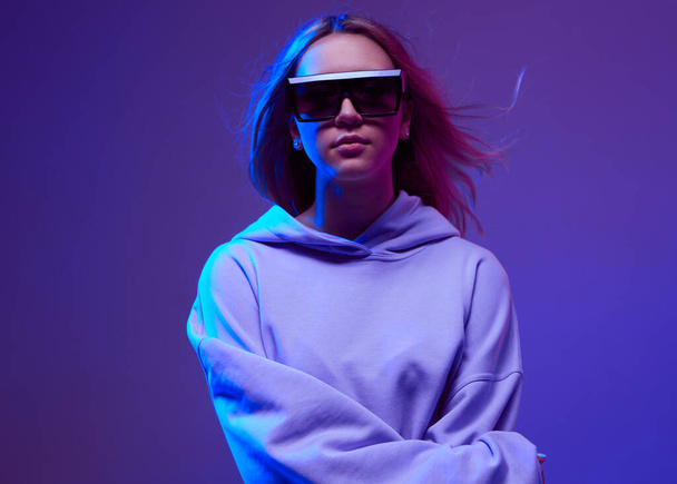 Portrait of a stylish young girl, cool posing in a hoodie, sunglasses and with developing hair, on a neon background. - Foto, afbeelding