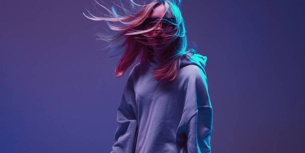 Portrait of a stylish young girl, cool posing in a hoodie, sunglasses and with developing hair, on a neon background. - 写真・画像