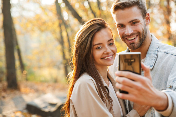 Beautiful young couple in love using mobile phone while spending time at the autumn park, taking a selfie - Фото, зображення