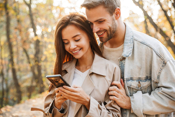 Beautiful young couple in love using mobile phone while spending time at the autumn park - Фото, изображение