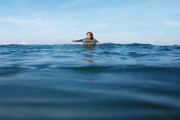 Surferem. Surfing człowiek na białej desce surfingowej Pływanie w oceanie. Sport wodny dla aktywnego stylu życia. Piękne morze i błękitne niebo na tle. - Zdjęcie, obraz