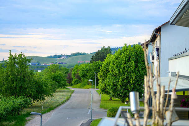 Árvore tranquila alinhada estrada rural em uma paisagem de primavera curvando casas passadas à direita em direção a colinas distantes
 - Foto, Imagem