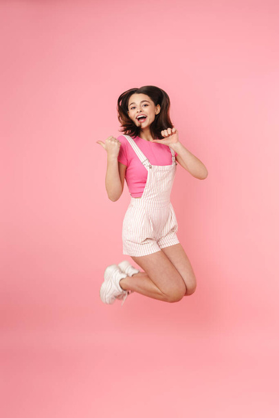 Full length of a pretty excited young teenage girl jumping isolated over pink background, rejoycing, pointing away - Foto, imagen