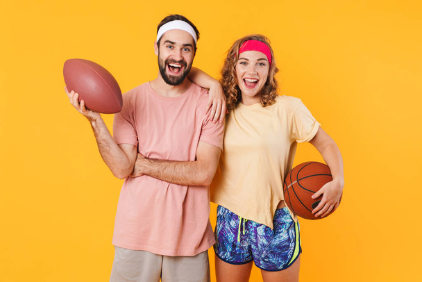 Portrait of young happy caucasian fitness couple wearing headbands holding sports balls isolated over yellow background - Фото, изображение
