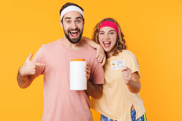 Portrait of young happy caucasian fitness couple wearing headbands holding protein jar and credit card isolated over yellow background - Foto, afbeelding