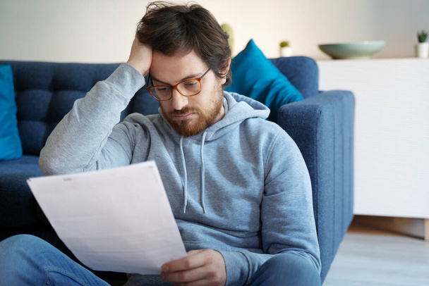 Ένας τύπος που διαβάζει αρνητικά νέα σε ένα γράμμα στο σπίτι στον καναπέ. - Φωτογραφία, εικόνα