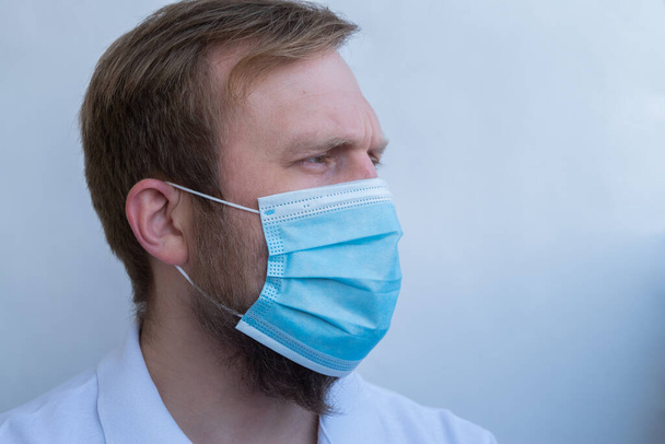 Men in a white t-shirt in a protective blue disposable mask on a white background close-up in profile. - Foto, Imagen