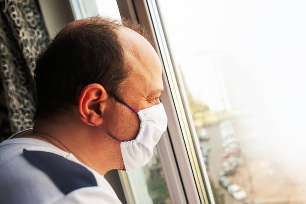 Mann mit medizinischer Schutzmaske schaut aus dem Fenster. Coronavirus-Konzept. Quarantäne-Konzept - Foto, Bild