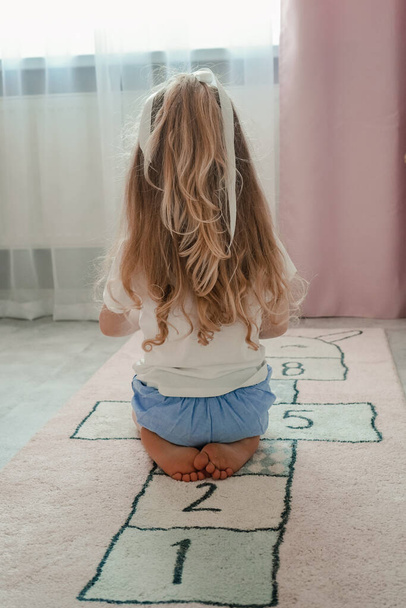 Malá holčička s dlouhými blond vlasy sedí se zády. Krásné a husté vlasy v dítěti. Blondýnka si hraje s hračkami v dětském pokoji. - Fotografie, Obrázek
