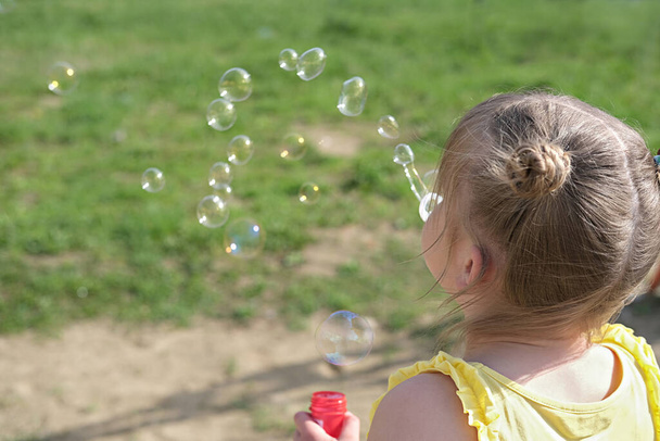 Mała dziewczynka robiąca bańki mydlane w letnim parku. słodka dziewczyna z bułki przestrzeni gra na świeżym powietrzu z baniek mydlanych. Koncepcja wakacji. widok z tyłu. - Zdjęcie, obraz